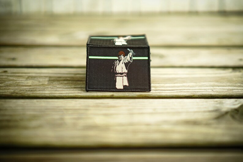 May include: A black and green embroidered patch featuring a character holding a lightsaber. The patch has a green line across the top and a white background.