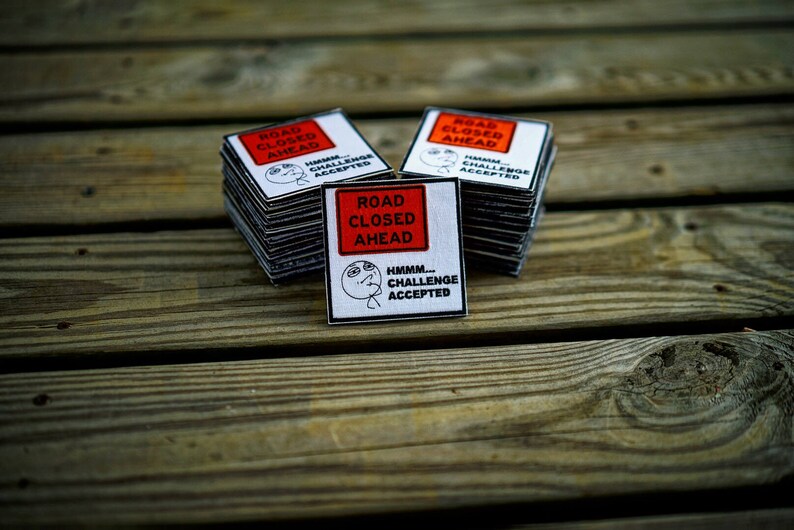 Op de afbeelding: Een stapel vierkante stickers in wit en rood met de tekst "Road Closed Ahead" en "Hmmm... Challenge Accepted".