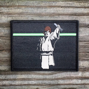 Op de afbeelding: Zwarte patch met een wit silhouet van een persoon die een lightsaber vasthoudt. De achtergrond is zwart met een groene lijn in het midden.