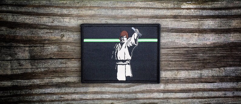 May include: Black patch with a white silhouette of a person holding a lightsaber. The background is black with a green line across the middle.