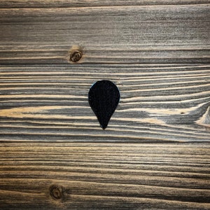 May include: A black fabric patch in the shape of a teardrop with a pointed tip. The patch is on a wooden surface.