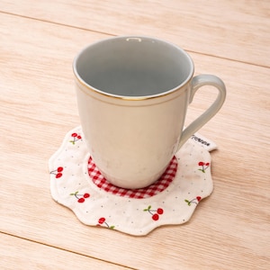 May include: A white ceramic mug with a gold rim sits on a cherry-patterned coaster. The coaster features a white fabric with red cherries and a red and white checkered center. The mug and coaster are on a light wood surface.