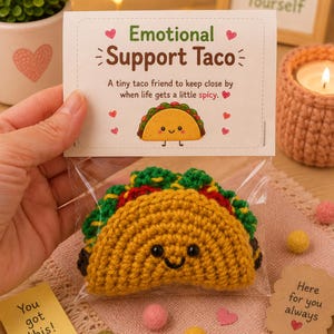 May include: A small, crocheted taco toy with a yellow shell, green lettuce, and red filling, featuring black bead eyes and a stitched smile. It is packaged in a clear plastic bag with a card labeled Emotional Support Taco with text.