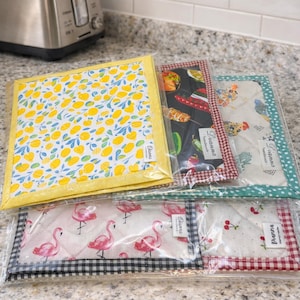 May include: A set of five square fabric pot holders in clear plastic packaging. Each features a unique print including yellow lemons, Mexican-themed motifs, chickens, pink flamingos, and red cherries, all with a small Dianna label.