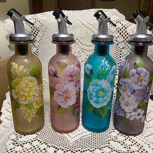 Puede incluir: Cuatro botellas de vidrio con diseños florales pintados a mano. Las botellas son en tonos de marrón, rosa, azul y morado. Cada botella tiene una tapa de bomba plateada.