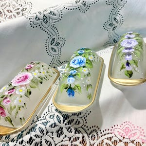 Op de afbeelding: Drie ovale glazen botervlootjes met goudkleurige metalen randen. Elk exemplaar is voorzien van handgeschilderde bloemmotieven: één met roze rozen, één met blauwe rozen en één met paarse bloemen, geaccentueerd met groene bladeren en kleine witte bloemetjes.