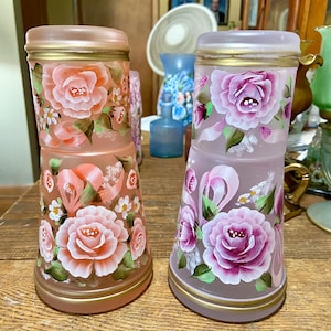 May include: Two glass tumblers with hand-painted floral designs. The tumblers are frosted glass with gold trim and feature pink and purple roses with green leaves and white ribbons.