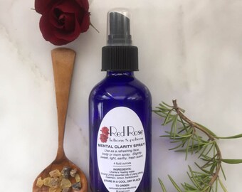 May include: A blue glass bottle with a black spray nozzle, labeled "Red Rose lotions & potions Mental Clarity Spray". The bottle is sitting on a white surface with a wooden spoon, a red rose, and a sprig of rosemary.