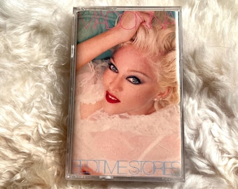 May include: A cassette tape with a pink and white design. The front of the tape features a photo of Madonna with the title "Bedtime Stories" printed in blue.