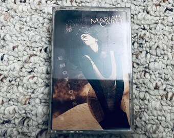 May include: A cassette tape of Mariah Carey's album "Emotions." The album cover features a dark image of Mariah Carey with her hair flowing. The word "Emotions" is vertically printed on the left side of the cover, and "Mariah Carey" is printed at the top.