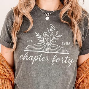May include: A gray t-shirt with the text "chapter forty" in a script font. Above the text is an open book with a floral design and the year "1985". The shirt is worn with a rust-colored cardigan and blue jeans.