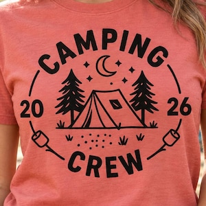 May include: A heathered coral short-sleeve t-shirt featuring a black graphic print of a tent, pine trees, a crescent moon, and stars. The text reads CAMPING CREW 2026, accompanied by illustrations of two marshmallows on sticks at the bottom.