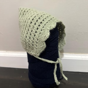 May include: A light sage green crocheted bonnet featuring a textured stitch pattern with a scalloped edge and thin matching ties. The bonnet is displayed resting on a dark navy blue ball of yarn to maintain its rounded shape and structure.