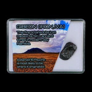May include: A dark, irregularly shaped meteorite specimen encased in a clear plastic display case. The case features a label with the text "Gibeon (Iron, IVA)" and information about its origin in Namibia. The background shows a landscape.