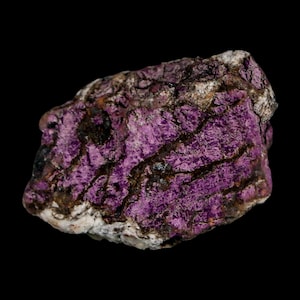 May include: A close-up of a rough-hewn, natural stone with a deep purple hue. The stone has a textured surface with dark brown and black veins running through it. The edges are a mix of white and brown, set against a black background.