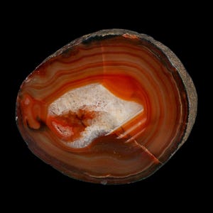 May include: A polished agate slice featuring concentric bands of deep orange, rust red, and brown hues. The center of the stone contains a crystalline cavity with white, translucent quartz formations, surrounded by a rough, dark outer edge.