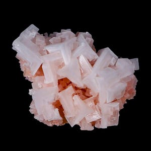 May include: A close-up of a cluster of pale pink mineral crystals against a black backdrop. The crystals are blocky and geometric, with sharp edges and a translucent quality. The overall shape is irregular and textured.