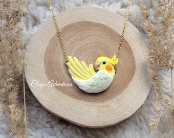 May include: A gold-colored chain necklace featuring a handcrafted pendant shaped like a yellow and white cockatiel bird. The bird pendant has detailed wings and a small orange beak. The necklace is displayed on a wooden slice.