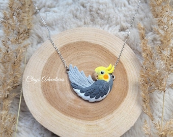 May include: A silver necklace featuring a handcrafted polymer clay pendant in the shape of a cockatiel bird. The bird has gray, white, and yellow coloring. The necklace is displayed on a wooden slice with the text "Clays Adventures".