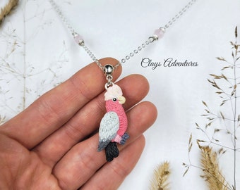 May include: A silver necklace featuring a pendant of a pink and gray bird. The bird pendant has a white crest and black tail feathers. The necklace also includes pink beads and the text "Clays Adventures".