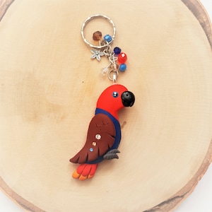 May include: A handmade parrot keychain featuring a red head, brown wings, and blue accents. The bird has a black beak and is adorned with small crystals. The keychain includes a silver ring and decorative beads.