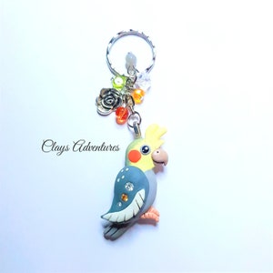 May include: A handmade parrot keychain with a silver keyring and decorative charms. The parrot is yellow, blue, and grey, with rhinestone accents. The keychain includes a silver rose charm and orange and clear beads. The text "Clays Adventures" is visible.