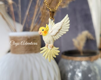 May include: A gold-colored chain necklace with a pendant featuring a detailed, cream-colored cockatiel with yellow and orange accents. The bird has outstretched wings and a detailed tail. The necklace is displayed against a blurred background.