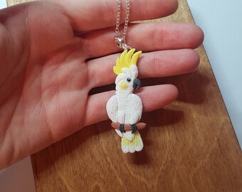 May include: A white and yellow cockatoo pendant necklace. The bird is perched on a brown branch, with a yellow crest and black beak. The necklace chain is silver. The pendant is held in a hand.