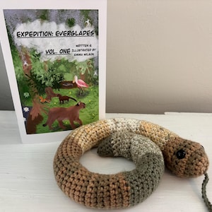 May include: A crochet snake toy featuring a variegated pattern of brown, tan, and olive green yarn with black button eyes and a thin green tongue, positioned in front of a book titled Expedition: Everglades Vol. One by Emma Wilson.