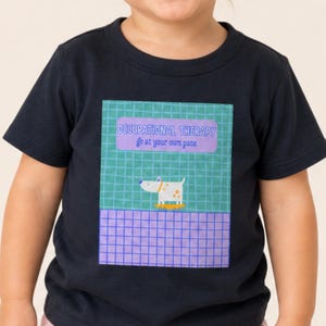 Pode incluir: Uma t-shirt de manga curta azul-marinho escuro com uma estampa gráfica retangular. O gráfico apresenta um fundo de grade verde-azulado, uma seção roxa com o texto "OCCUPATIONAL THERAPY go at your own pace" e um pequeno cão branco sobre uma base amarela.
