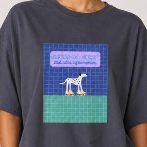 Pode incluir: Uma t-shirt cinzento-carvão com uma impressão gráfica de um cão dálmata branco sobre patins. O design inclui um fundo de grelha azul e uma faixa retangular roxa com o texto "OCCUPATIONAL THERAPY small wins, big momentum."