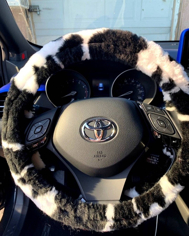 May include: Black and white cow print steering wheel cover made of soft, fuzzy material. The cover is installed on a black steering wheel with a silver Toyota logo and the words "SRS Airbag".