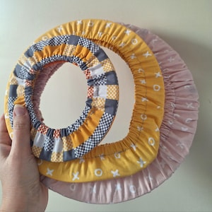 以下が含まれることがあります： さまざまなサイズの布で覆われた3つのフープ。上部のフープは、グレー、白、黄色のパッチワークデザインです。中央のフープは、白い「x」と「o」のパターンが入った黄色です。下のフープは薄いピンク色です。