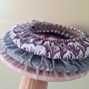 May include: Three fabric bowl covers stacked on top of each other. The top cover has a white and purple floral pattern. The middle cover is grey with white dots. The bottom cover is pink with white crosses and circles.