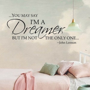May include: A black and white wall decal with a quote from John Lennon: "...You may say I'm a Dreamer, but I'm not the only one..." The quote is displayed on a light beige wall with a white bed and pillows in the background.