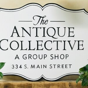 Könnte beinhalten: Ein weißes Schild mit schwarzem Rand und Text "The Antique Collective A Group Shop 334 S. Main Street". Das Schild hat einen dekorativen, wellenförmigen Rand.