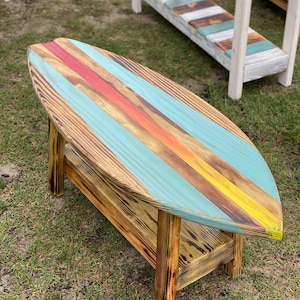 May include: A surfboard-shaped coffee table with a distressed wood finish. The top is painted with blue, yellow, orange, and red stripes. The table has a lower shelf.