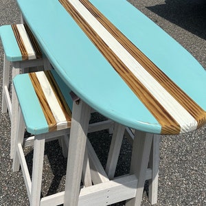 May include: A surfboard-shaped table with a light blue top and brown and white stripes. Two white wooden stools with light blue seats and brown and white stripes are included.