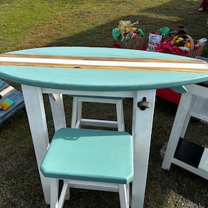 May include: A teal surfboard-shaped table with a white base and a teal stool. The table has a white stripe down the center and a bottle opener on the side.