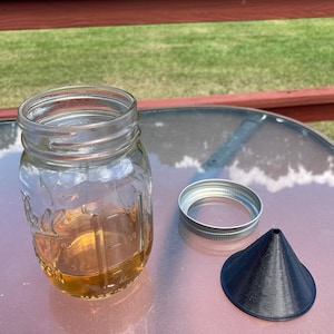 May include: A clear glass jar with a metal lid and a black plastic funnel. The jar is filled with a light brown liquid.