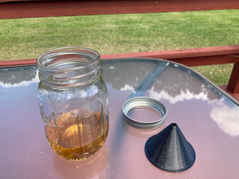May include: A clear glass jar with a metal lid and a black plastic funnel. The jar is filled with a light brown liquid.