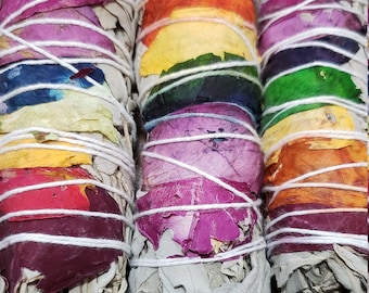 May include: Three white sage smudge sticks wrapped with white string and decorated with colorful flower petals. The petals are arranged in a rainbow pattern.