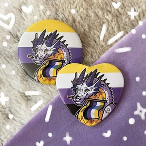 May include: Two button pins featuring a dragon with a purple, white, and yellow flag. The dragon is on a yellow, white, and purple background. The pins are heart-shaped and round.
