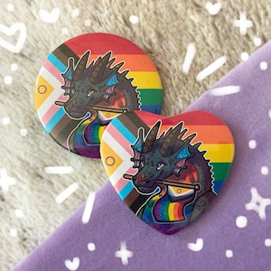 May include: Two round pinback buttons featuring a black dragon with rainbow wings and a pride flag design. The buttons are on a purple background.
