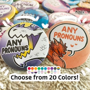 May include: Two button pins with the text "ANY PRONOUNS" in a comic book style. The left pin features a white dragon with purple and yellow accents. The right pin features an orange dragon with a speech bubble. The text "Choose from 20 Colors!" is below the pins.