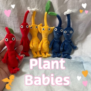 May include: A collection of six plush figures in red, yellow, and blue, each featuring large white eyes, a slender stem-like head appendage, and a small flower or leaf topper. The text "Plant Babies" is displayed in white with a pink outline.