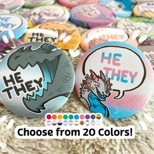May include: Two 1.25 inch button pins with a dragon and a speech bubble. The dragon is gray and blue. The speech bubble is pink and blue with the text "HE THEY" in white. The buttons are surrounded by other buttons with various colors and designs.
