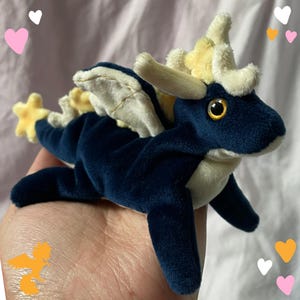May include: A plush dragon toy in navy blue with cream-colored accents, including wings and horns. The dragon has a yellow eye and a star-shaped tail. The toy is made of soft material.