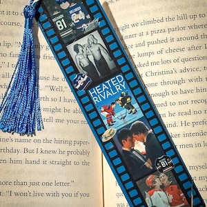 May include: A blue bookmark with a film strip design featuring hockey-themed images and the text "HEATED RIVALRY." It has a blue tassel attached. The bookmark is placed on top of an open book.