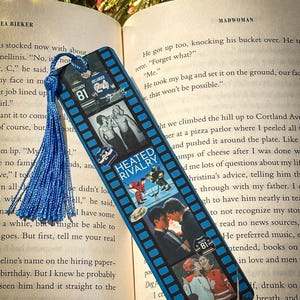 May include: A blue bookmark with a film strip design featuring hockey-themed images and the text "HEATED RIVALRY." The bookmark has a blue tassel and is resting on the pages of a book.